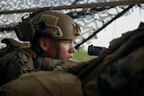 A close-up of a soldier in a military helmet peering through a rifle's scope.