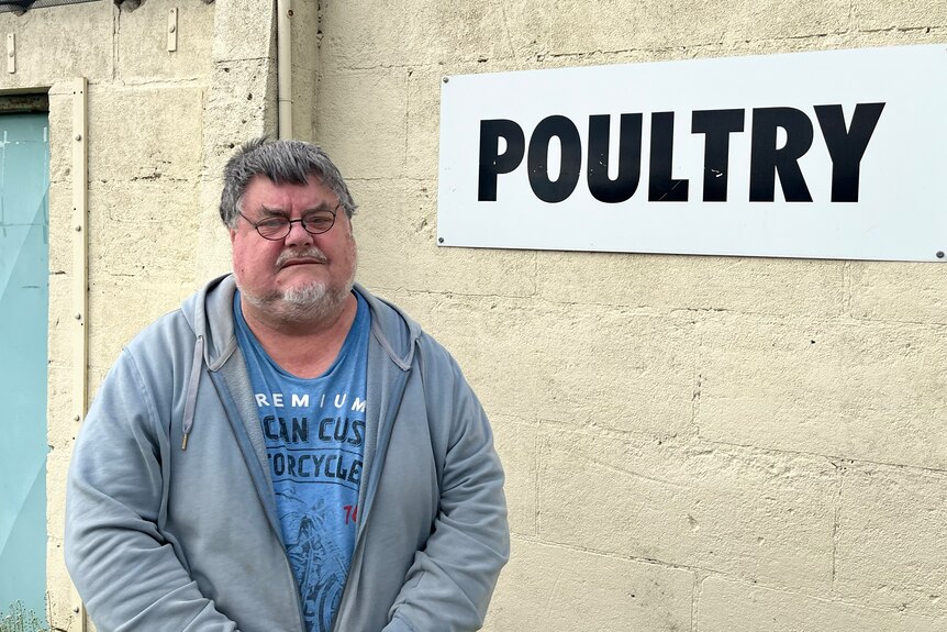 Large seriuous man, greying hair, beard, blue tee, hoodie, standing in front of a sign on a brick wall that says poultry.