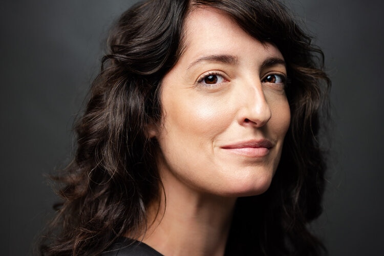 Rachel Yoder, with long wavy brown hair and dark shirt, stands against grey background smiling.