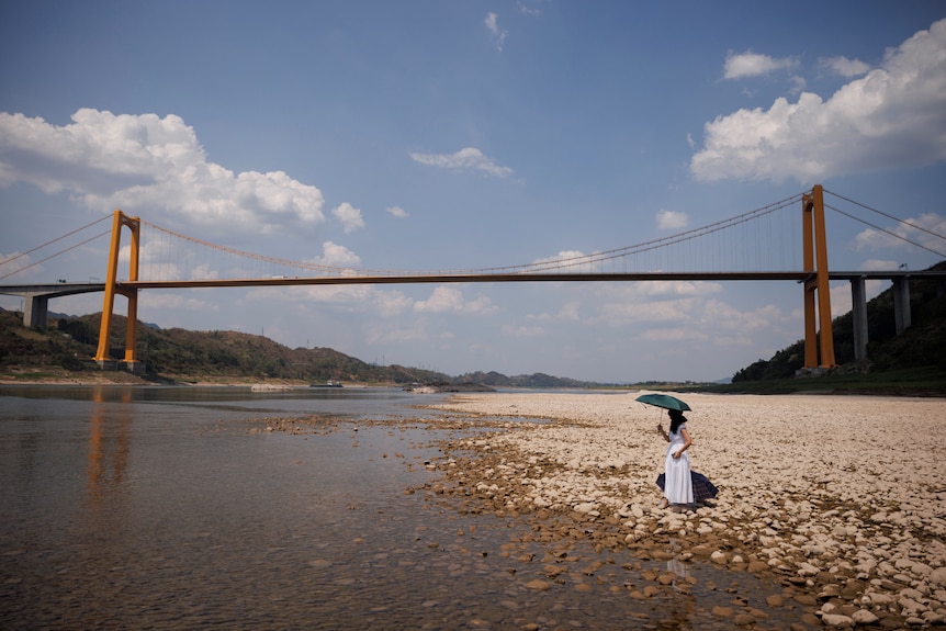 A woman holding an umbrella stands on a rocky shoreline next to a shallow river. A suspension bridge spans the horizon