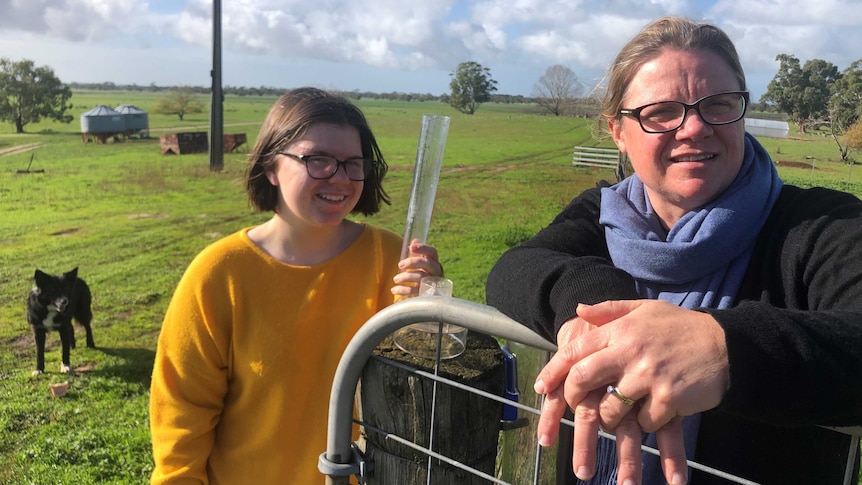 A woman wearing a blue scarf alongside a girl in a yellow jumper stand by a gate in a paddock.