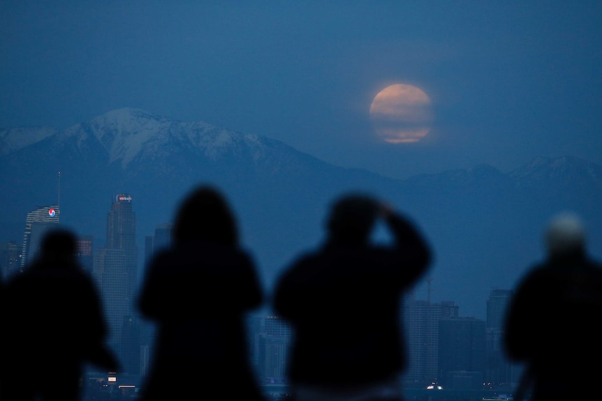 The reddish-hued moon is visible behind some clouds above the mountain range.