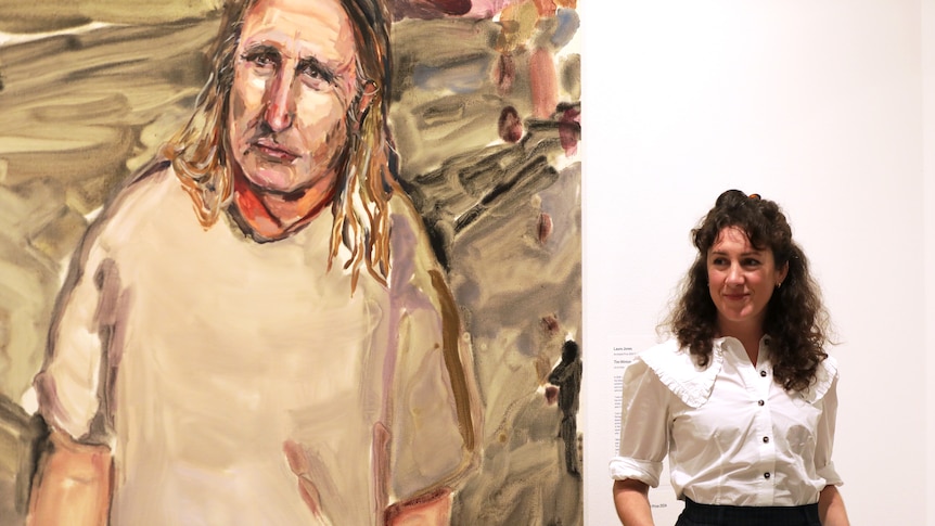 A woman in her early 40s - the artist Laura Jones - smiling and standing in front of her painting of Tim Winton