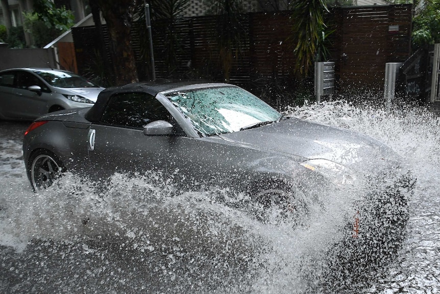 Flood water splashes up around a grey car as it drives down flooded Melbourne street.