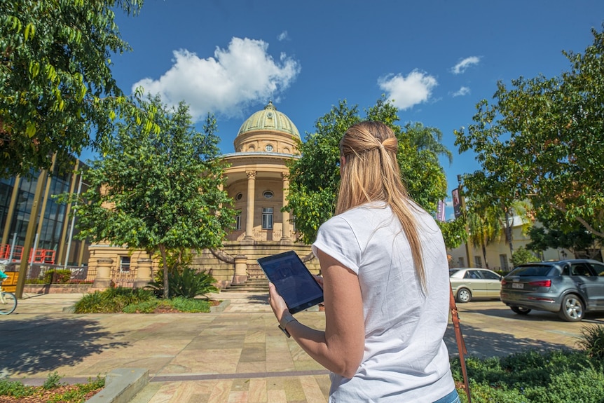 Woman stands in front of Customs House in Rockhampton, holding an tablet device displaying the app.