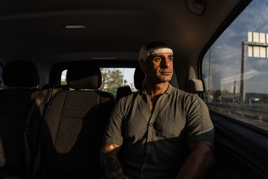 A man wearing a white bandage around his head looks out a car window as the setting sun beats down on him