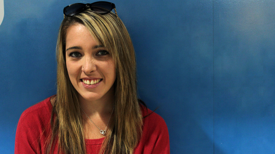 A woman with blonde hair, sunglasses and a red sweater standing in front of a blue background.