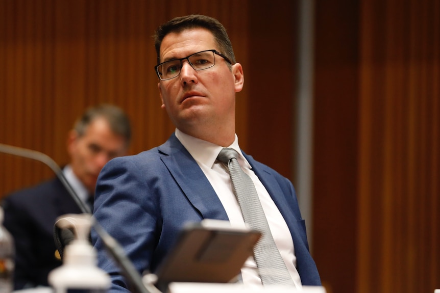 Politician Zed Seselja sits in a room at a table, wearing a blue suit and square glasses.