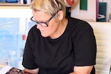 A woman dressed in black, with short blonde hair and glasses, focusses while needlepointing, in a room full of coloured thread