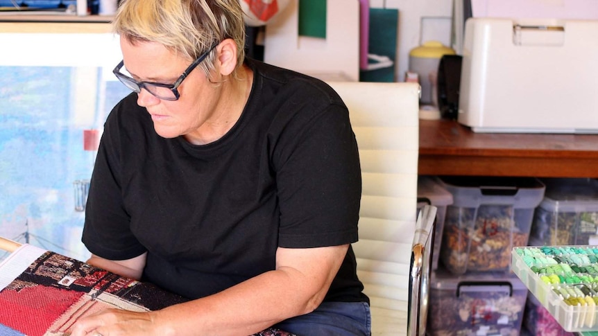 A woman dressed in black, with short blonde hair and glasses, focusses while needlepointing, in a room full of coloured thread