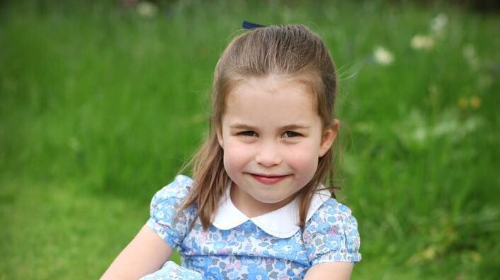 Princess Charlotte smiles wearing a blue floral dress as she sits on the grass with her arms wrapped around her knees