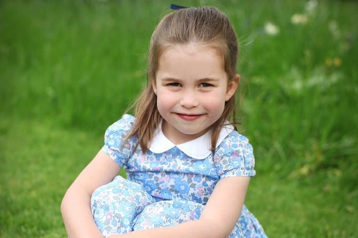 Princess Charlotte smiles wearing a blue floral dress as she sits on the grass with her arms wrapped around her knees