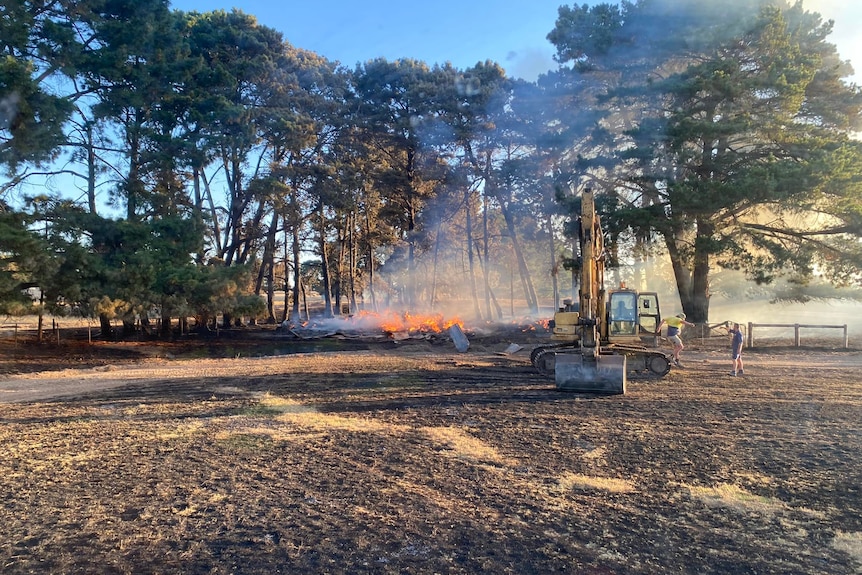 A small fire burns in a burnt paddock with digging machinery next to it, trees around.