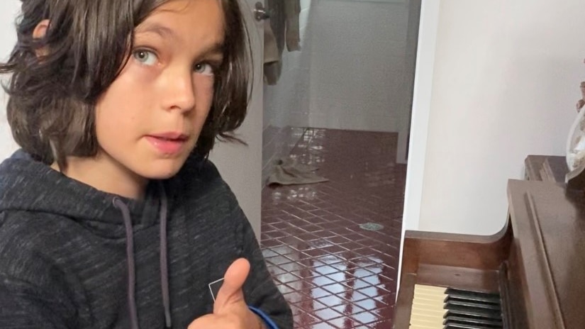 A young boy with shoulder-length brown hair sitting in front of an upright piano giving 'thumbs up'.