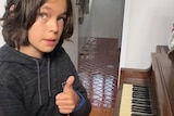 A young boy with shoulder-length brown hair sitting in front of an upright piano giving 'thumbs up'.