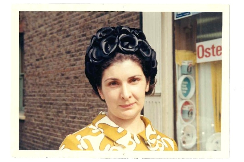 A young woman wearing a large black hat and a yellow and white buttoned frock stands in front of a shop window.