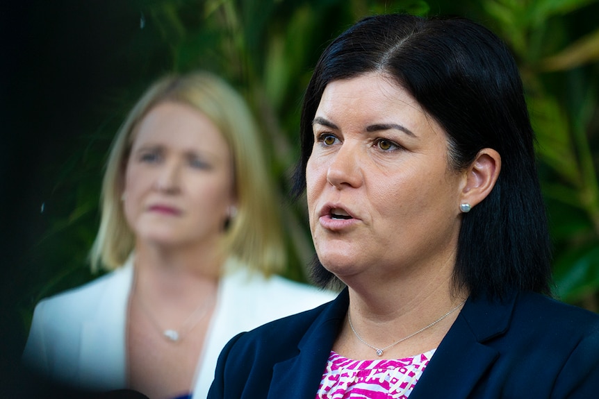 A woman with short dark hair speaks, behind her is a woman with short blonde hair. They both look serious.