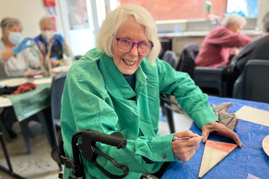 Older woman in a green overcoat smiles down the camera whilst using water colour paint