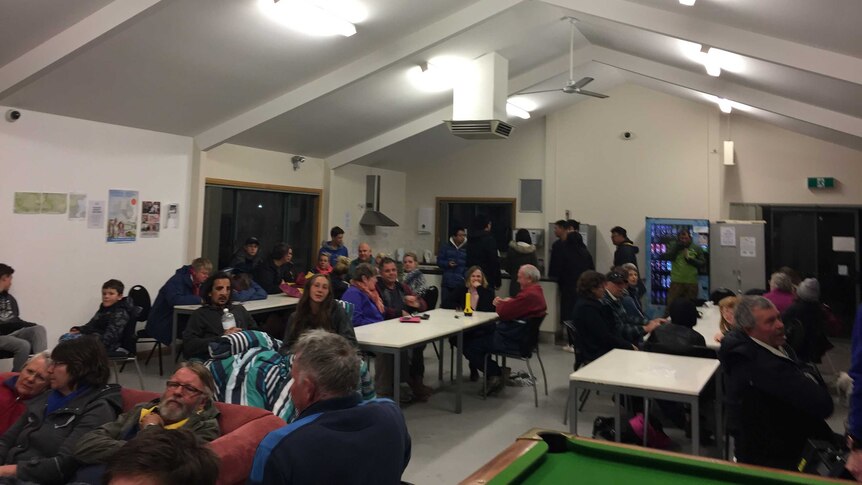 A large group of evacuated campers sit around a room talking, resting, eating.