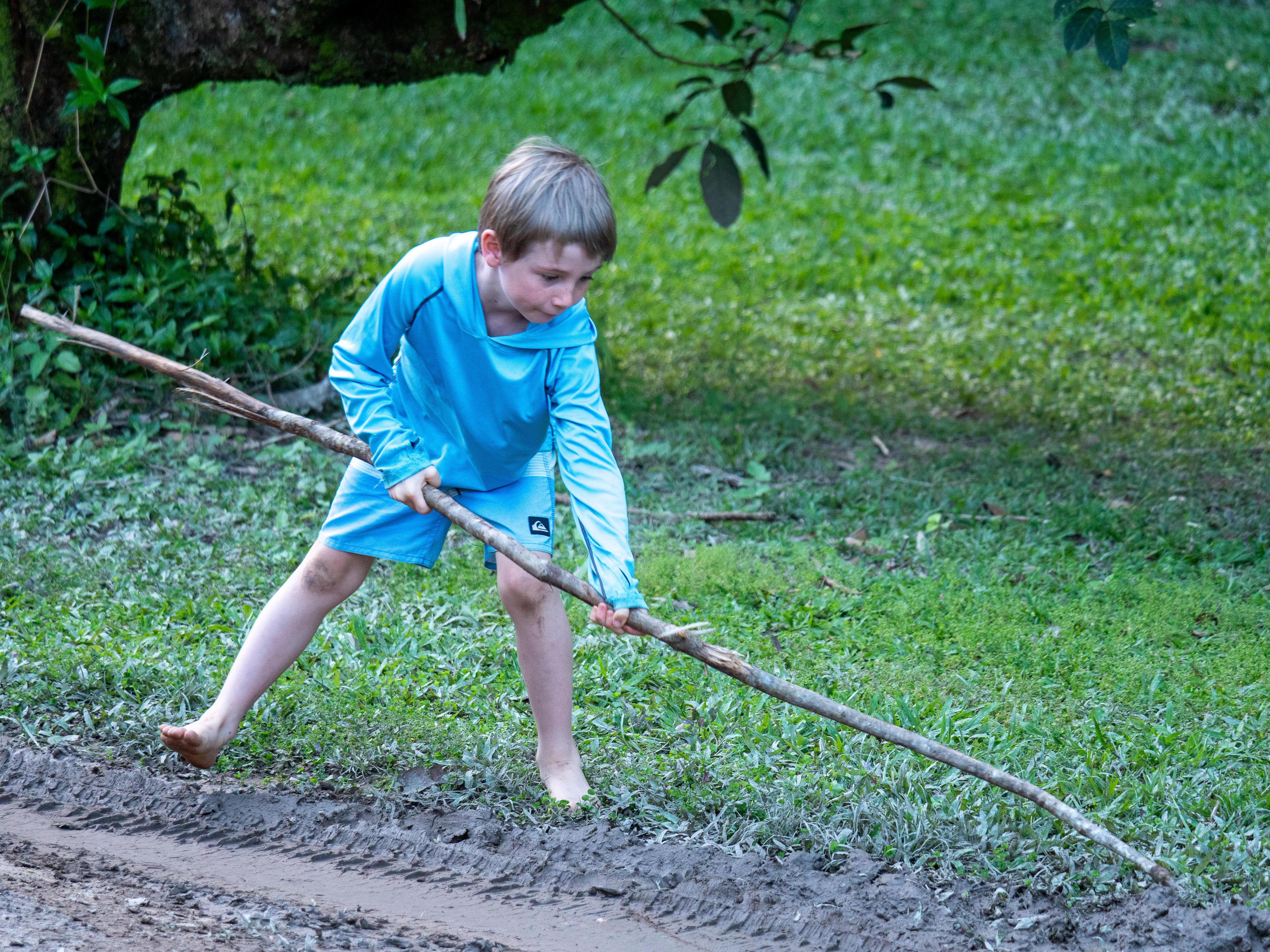 Boy wearing blue pushing a stick into some mud in a lush setting.