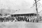 A black and white photo with about 100 people standing in front of an old country pub.