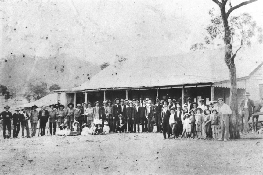 A black and white photo with about 100 people standing in front of an old country pub.