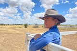 A woman in a work shirt and broad-brimmed hat leans on a fence, looking out over a rural property.