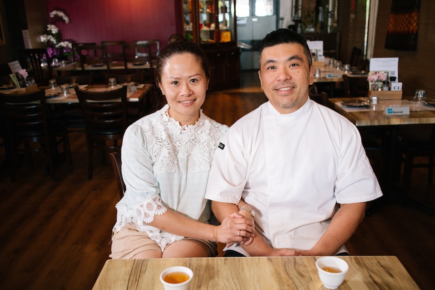 A woman holds the arm of a man wearing a white chef's top as they sit at a small square table in an empty restaurant.