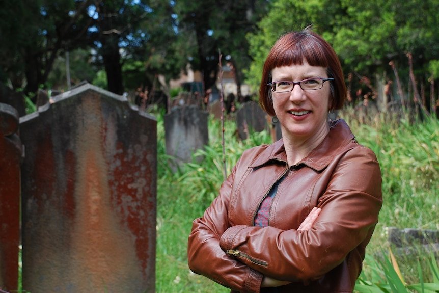 Dr Lisa Murray stands beside a old headstone, in a cemetery that resembles a park with longer, unkept grass