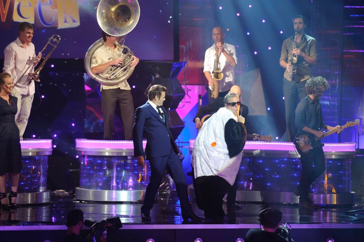 Μια τηλεόραση με τον Mikey Robins, 40, με κοστούμι και γυαλιά ηλίου σε στυλ πιγκουίνου, με τον Paul McDermott, ένα ζωντανό συγκρότημα να παίζει πίσω.