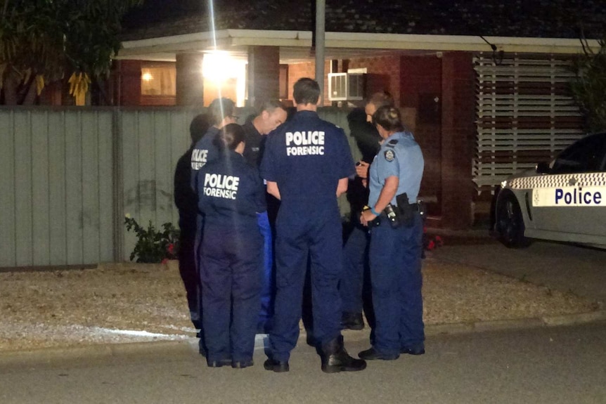 Police stand in a group outside the home. A police car is in the driveway.