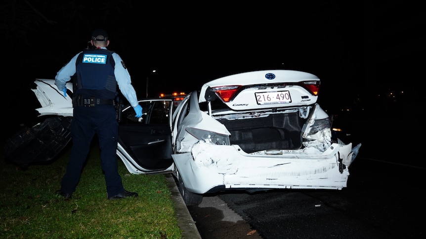 A police officer stands next to a smashed white sedan.