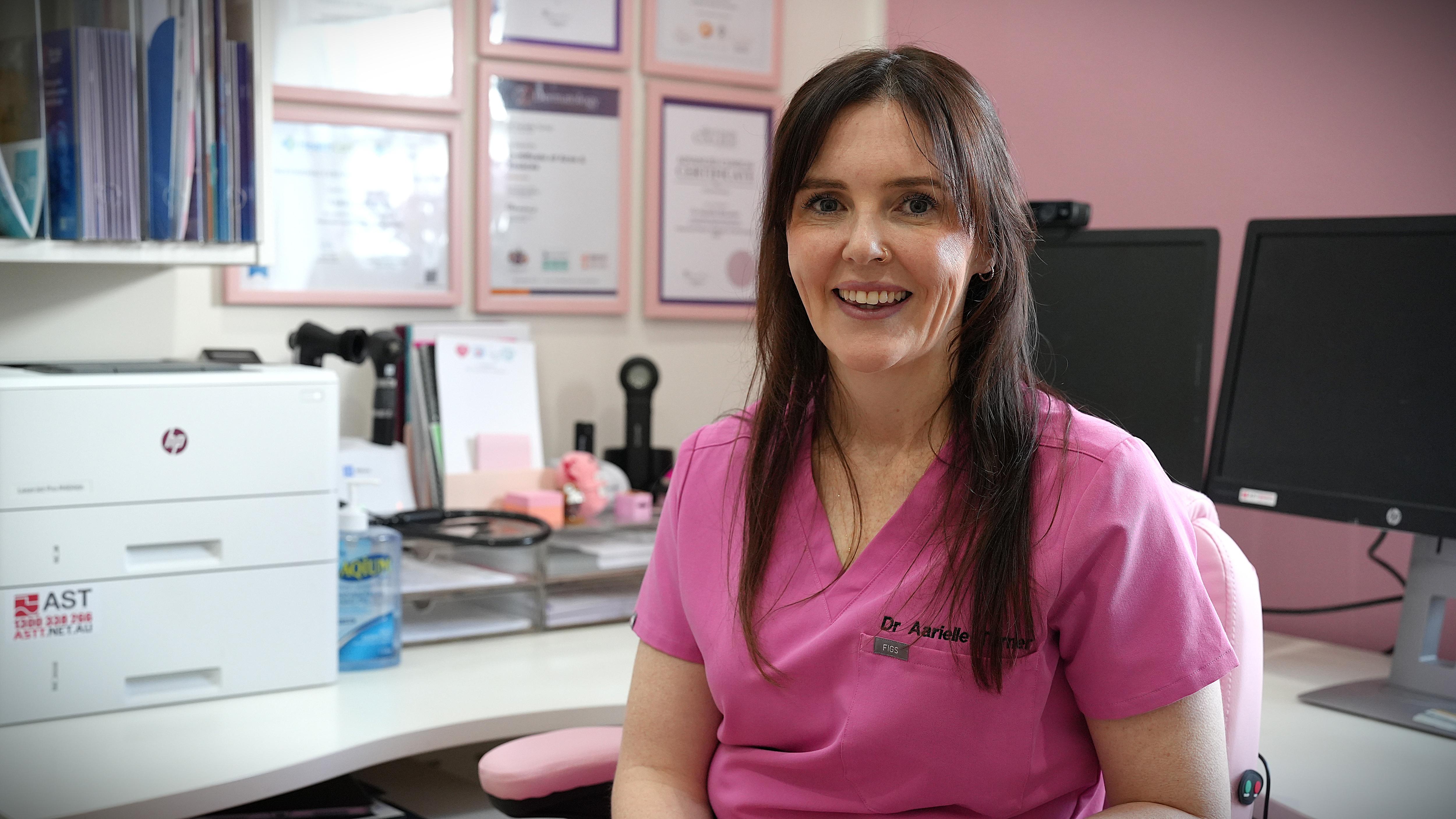 A smiling, dark-haired woman in pink scrubs sitting inside a pink-painted GP's office.