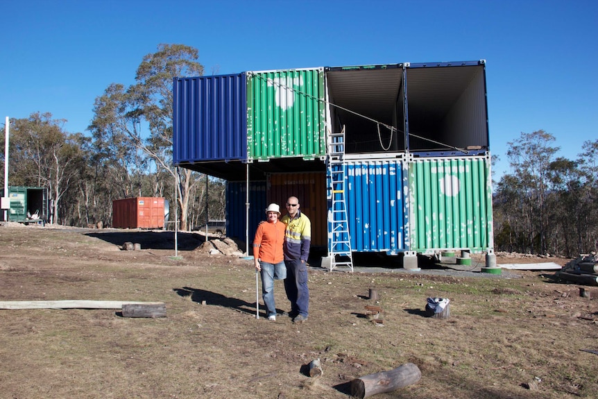 Two people stand in front of a shipping container house under construction.