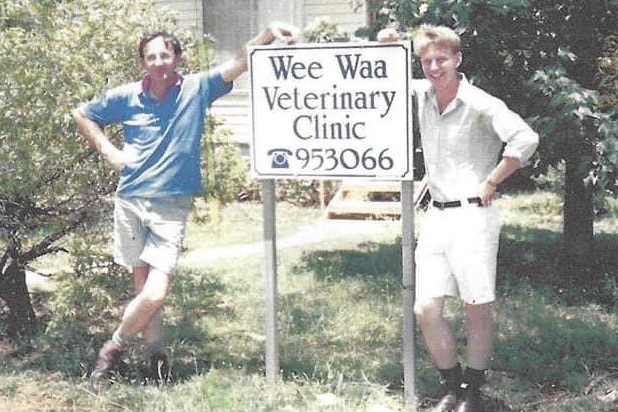 Two men lean on a sign out the front of a small wooden building. They are smiling