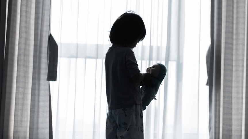 a generic image of a child standing by a window holding a doll. the light is shadowy and their identity isn't clear