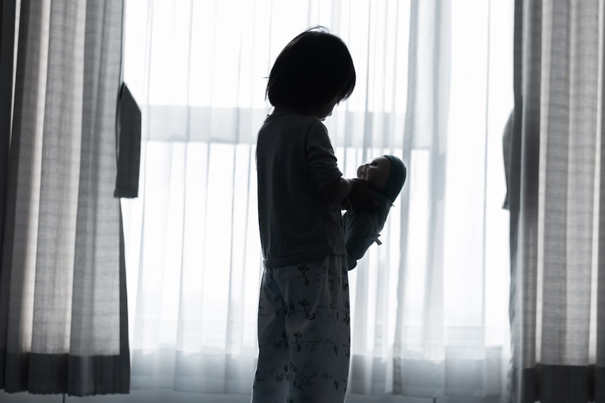 a generic image of a child standing by a window holding a doll. the light is shadowy and their identity isn't clear