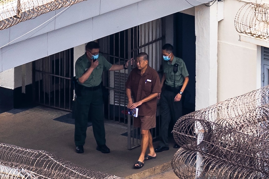 Jimmy Lai wearing brown shorts and shirt walks through a prison escorted by two guards.