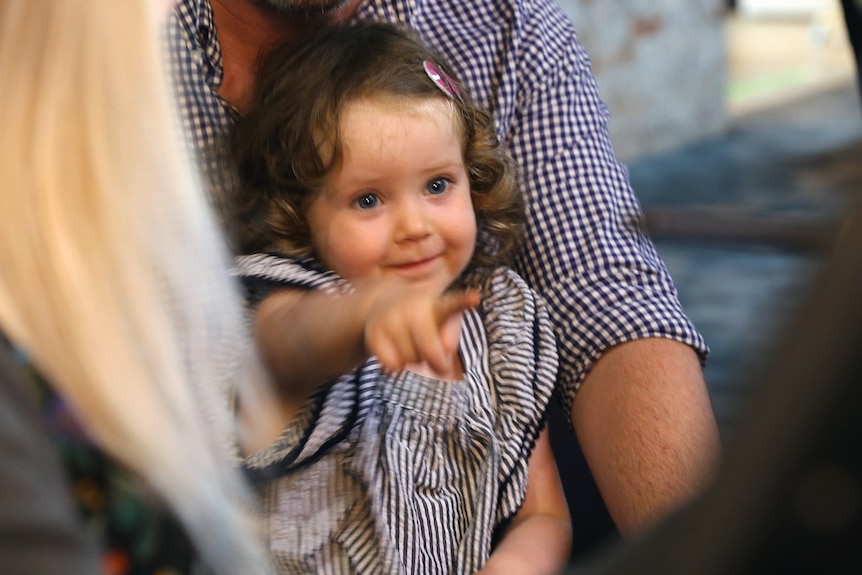 A girl sitting on her dad's lap and pointing at a screen and smiling.