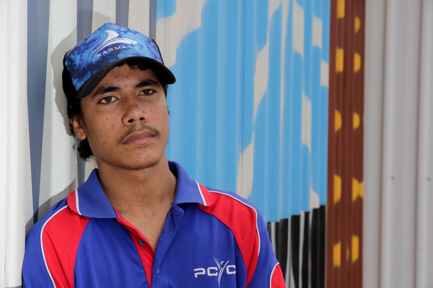 A young Indigenous man in a PCYC polo shirt and hat leans on the muralled wall of a corrugated steel building.