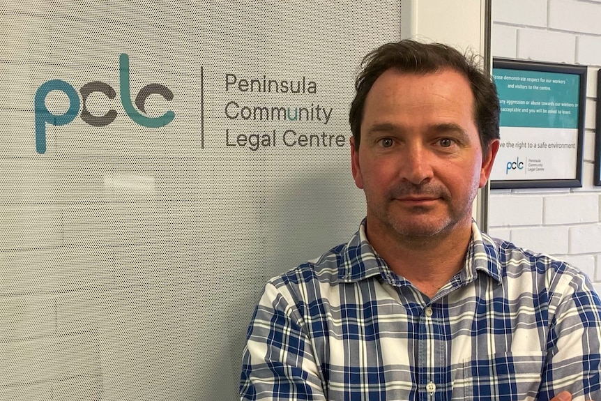 A man in a checked blue shirt with arms crossed in front of a peninsula community legal centre sign.