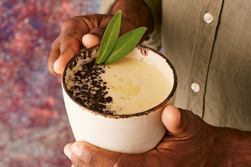 Damien is holding a latte in a white coffee cup with chocolate sprinkled on top and two pepperberry leaves