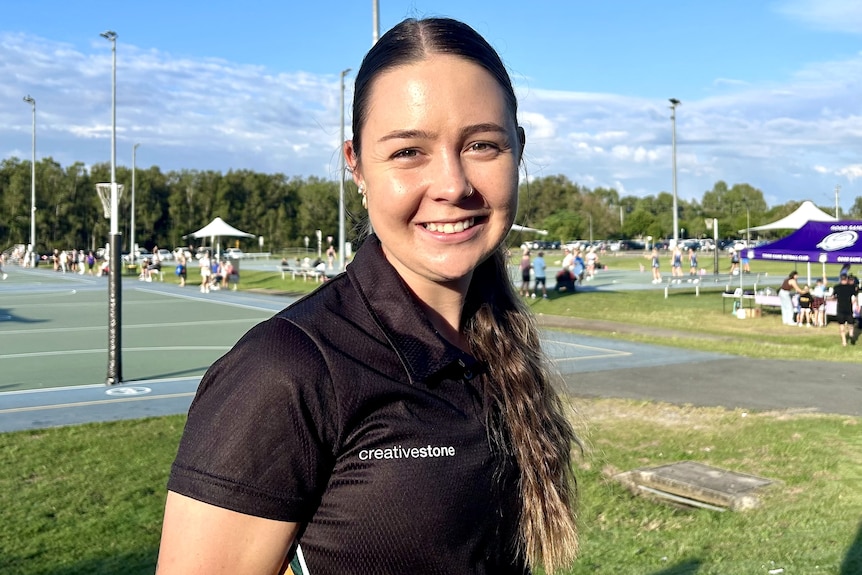 A young woman with long dark hair, tied back, wearing a dark shirt. She is standing near a netball court, smiling.