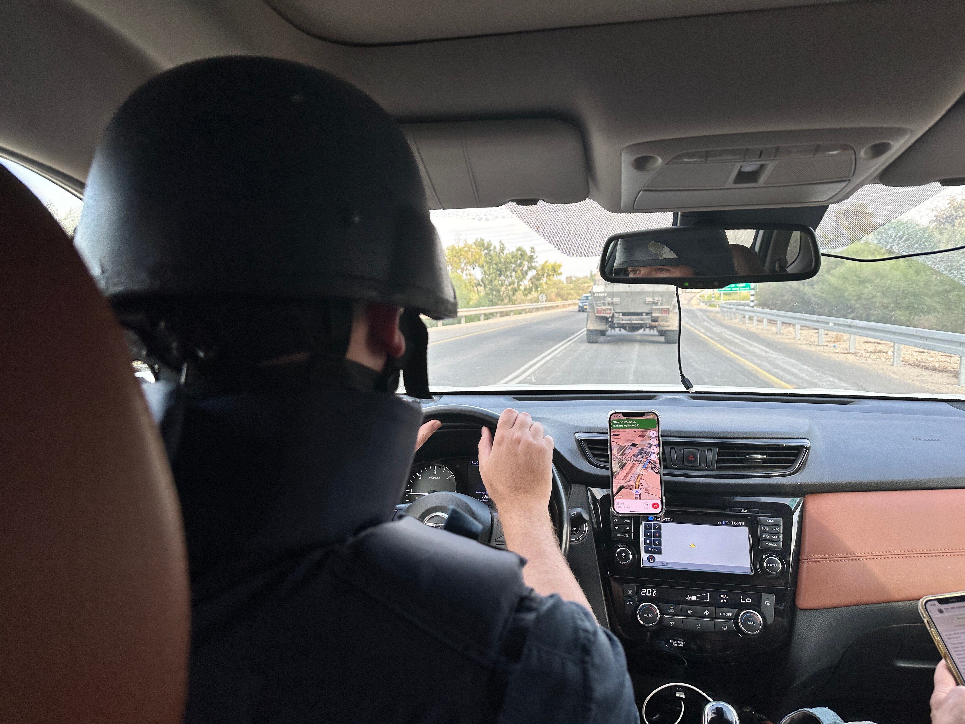 Point of view of a man wearing flak jacket and helmet driving a car with an armoured vehicle ahead on road.