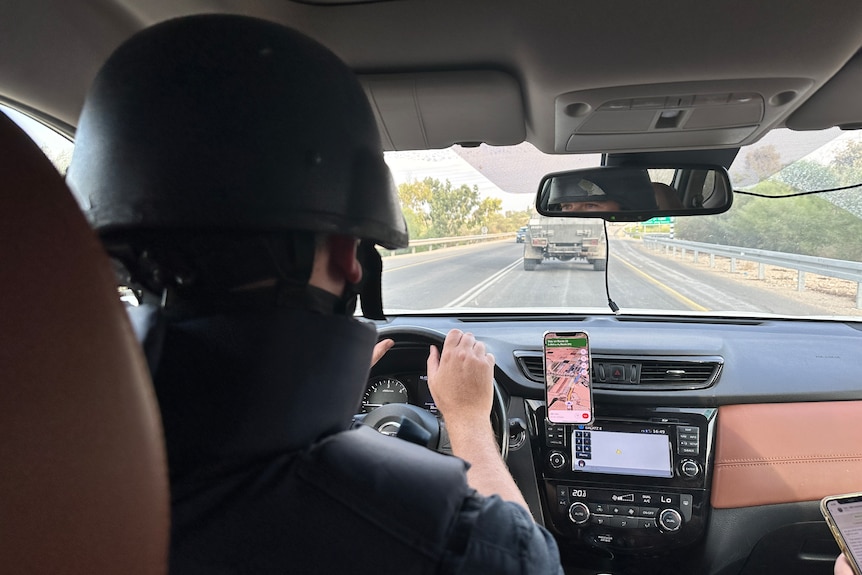 Point of view of a man wearing flak jacket and helmet driving a car with an armoured vehicle ahead on road.