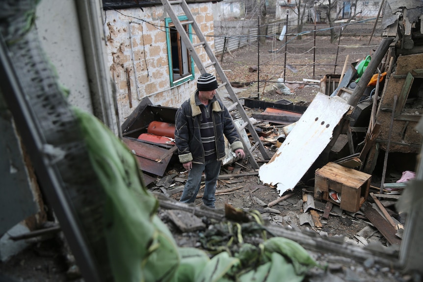 A man stands among the debris of his destroyed house, examining the damage.