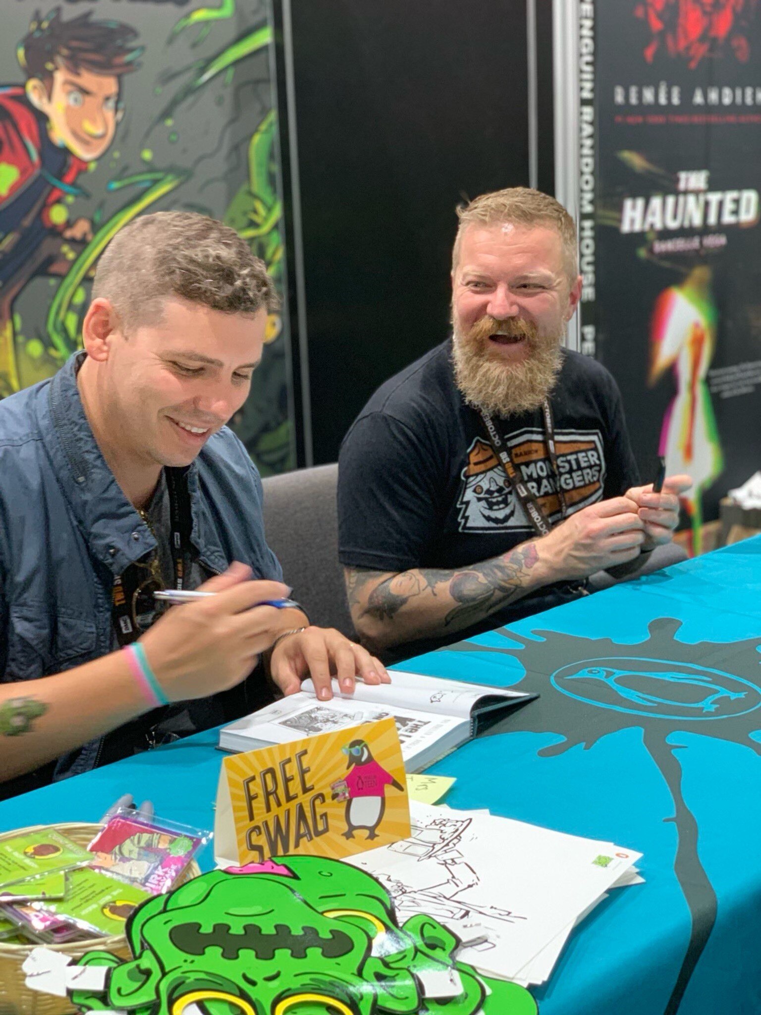 Two guys sit at a table signing graphic novels, smiling. 