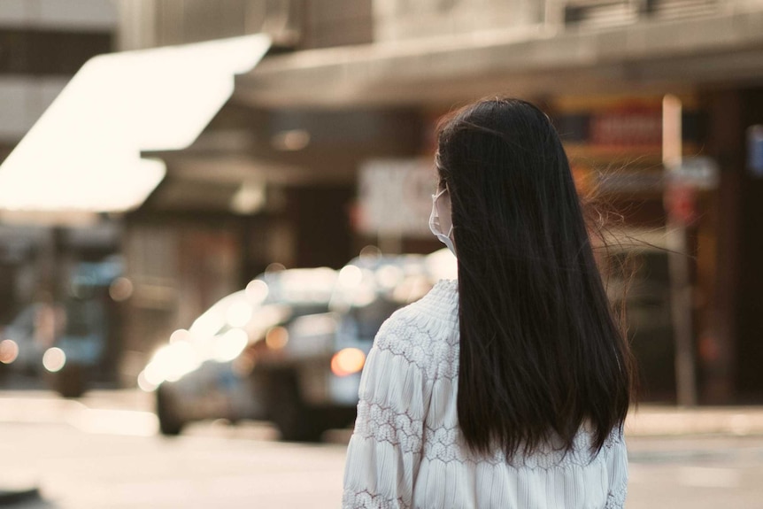 A woman in a mask stands on a street corner.