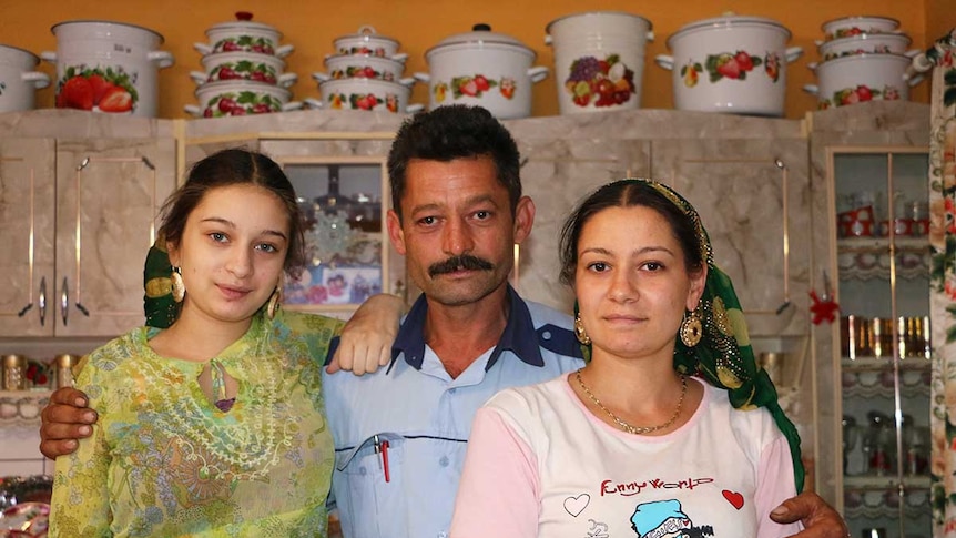Two sisters stand beside their father in their home kitchen.