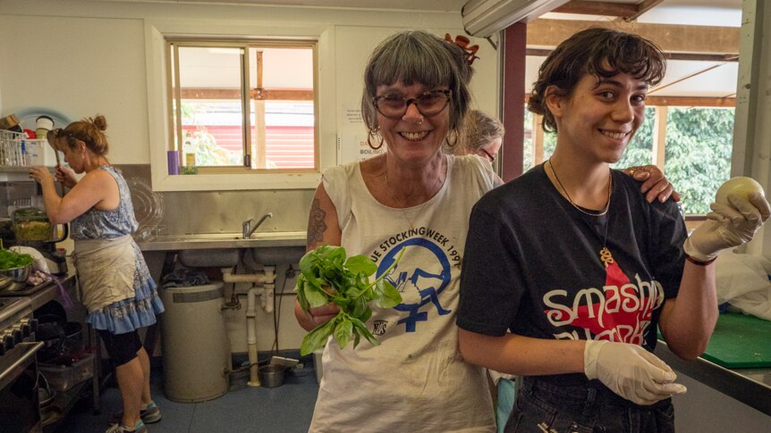 Two woman smiling at camera, standing in a kitchen in a hall, one holding basil and the other an onion.